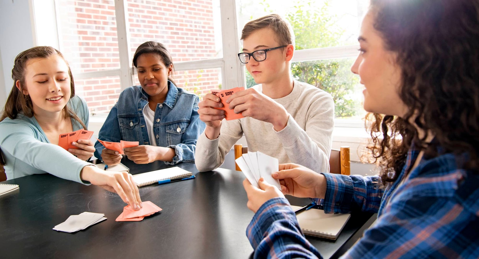 A group of four middle school age students sitting around a table. They are each holding some playing cards in their hands. There are piles of cards on the table in front of the students. One student has a pencil and notebook next to them. A group of four middle school age students sitting around a table. They are each holding some playing cards in their hands. There are piles of cards on the table in front of the students. One student has a pencil and notebook next to them.