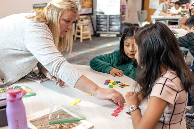 Elementary teacher guiding two students in a Eureka Math² lesson using colorful geometric tiles at a classroom table.