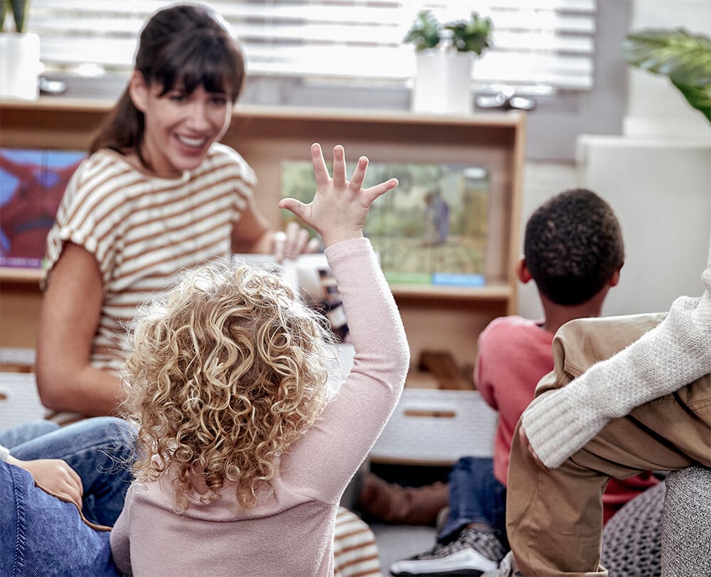 Student raising their hand as an educator reads them a Geodes book Student raising their hand as an educator reads them a Geodes book