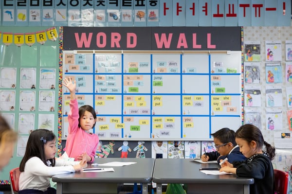 Four elementary students seated around a classroom table with one child raising her hand with a "Word Wall" on the board in the background.