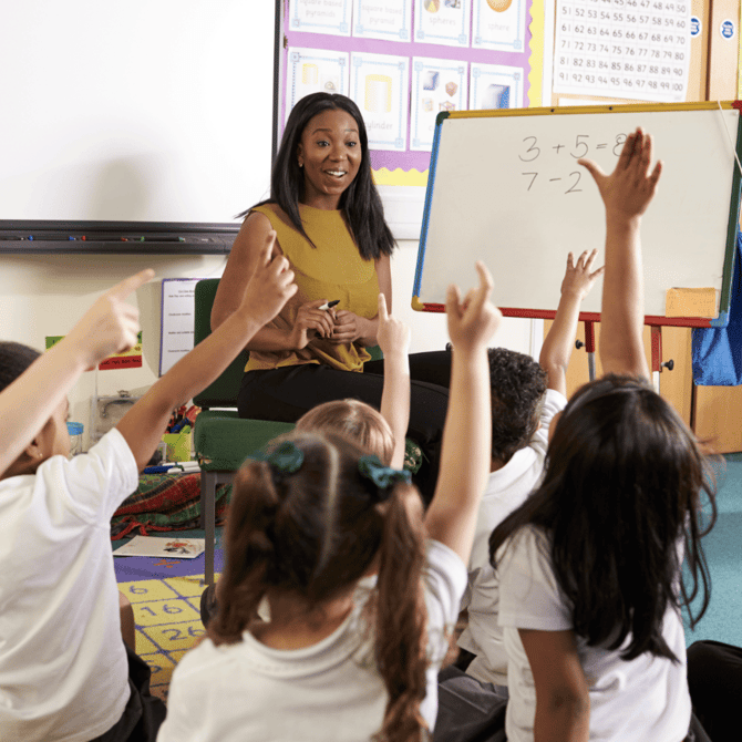 Students sitting on the carpet in front of their teacher. Students raise hands as the teachers writes number sentences on the whiteboard.