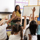 Students sitting on the carpet in front of their teacher. Students raise hands as the teachers writes number sentences on the whiteboard.